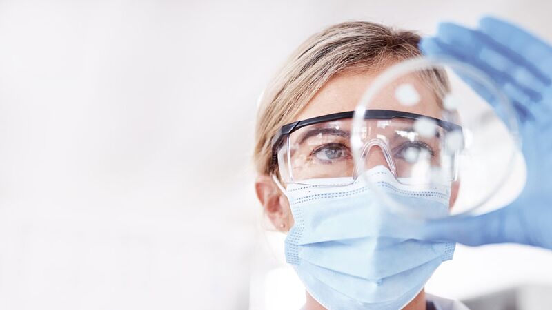 Eine Wissenschaftlerin mit Schutzbrille und Maske untersucht konzentriert eine Petrischale im Labor.
