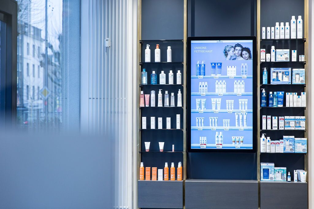 Ein Regal in einer Apotheke zeigt eine Auswahl an Hautpflegeprodukten für unreine, fettige Haut neben einem digitalen Display.