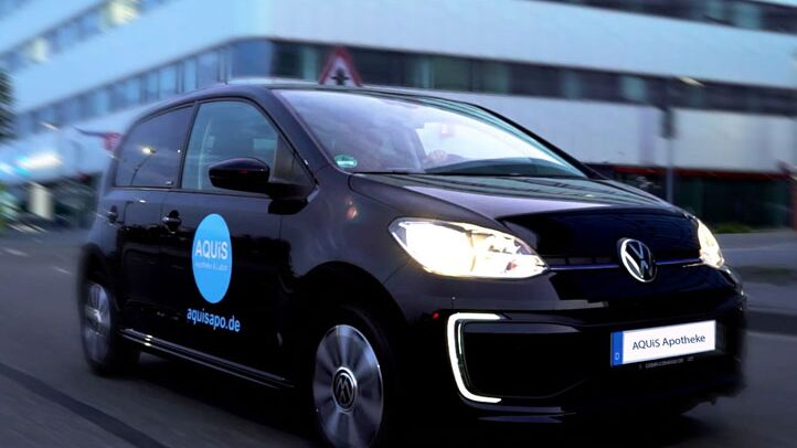 Ein schwarzes Auto mit "AQUIS Apotheke"-Logo fährt bei Dämmerung schnell durch eine städtische Straße.